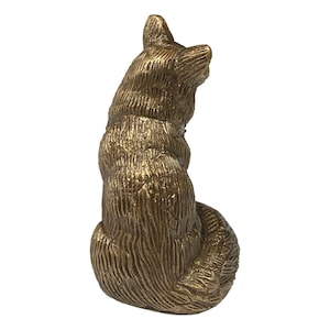 May include: A gold-colored metal figurine of a fox sitting with its tail curled around its body. The figurine is detailed with fur texture and has a realistic appearance.