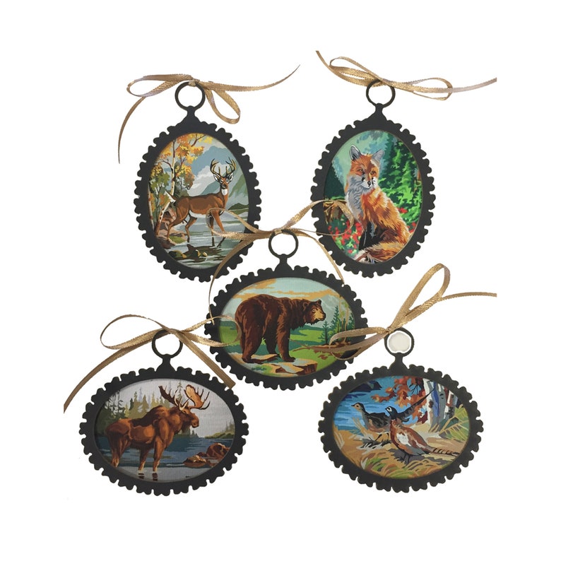 May include: Five oval-shaped ornaments with black metal frames and brown ribbon hangers. Each ornament features a different woodland animal: a deer, a fox, a bear, a moose, and a pair of pheasants.