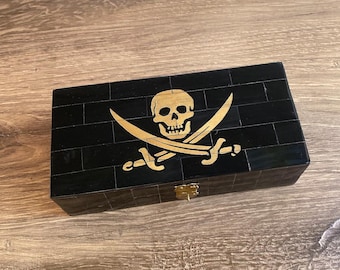 Puede incluir: Una caja de madera negra con un diseño de calavera y huesos cruzados dorados. La caja tiene un pequeño pestillo de latón.