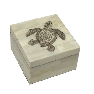 Könnte beinhalten: Eine weiße quadratische Box mit einem geschnitzten Meeresschildkröten-Design auf dem Deckel. Die Box ist aus Knochen und hat eine natürliche, cremefarbene Farbe.