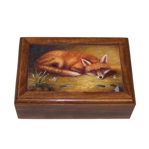 Pode incluir: Uma caixa de madeira com tampa articulada apresenta uma imagem pintada de uma raposa vermelha deitada sobre um fundo dourado com folhas espalhadas. A raposa tem um peito branco e uma cauda espessa.