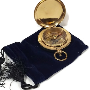 May include: A gold compass with a blue needle and a black and white compass rose. The compass is open and sitting in a blue velvet pouch with a black tassel.
