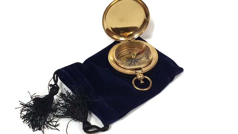 May include: A gold compass with a blue needle and a black and white compass rose. The compass is open and sitting in a blue velvet pouch with a black tassel.