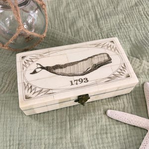 May include: A rectangular off-white box constructed from small tiled segments, featuring a dark brown etched illustration of a whale centered within an oval frame. The number 1793 is printed below the whale. A small brass-colored metal latch secures the lid.