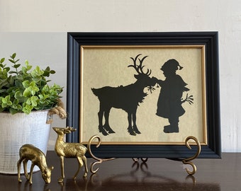May include: A black silhouette of a child feeding a reindeer, framed in a black frame with a gold mat. The frame is on a gold stand and there are two gold deer figurines in the foreground.