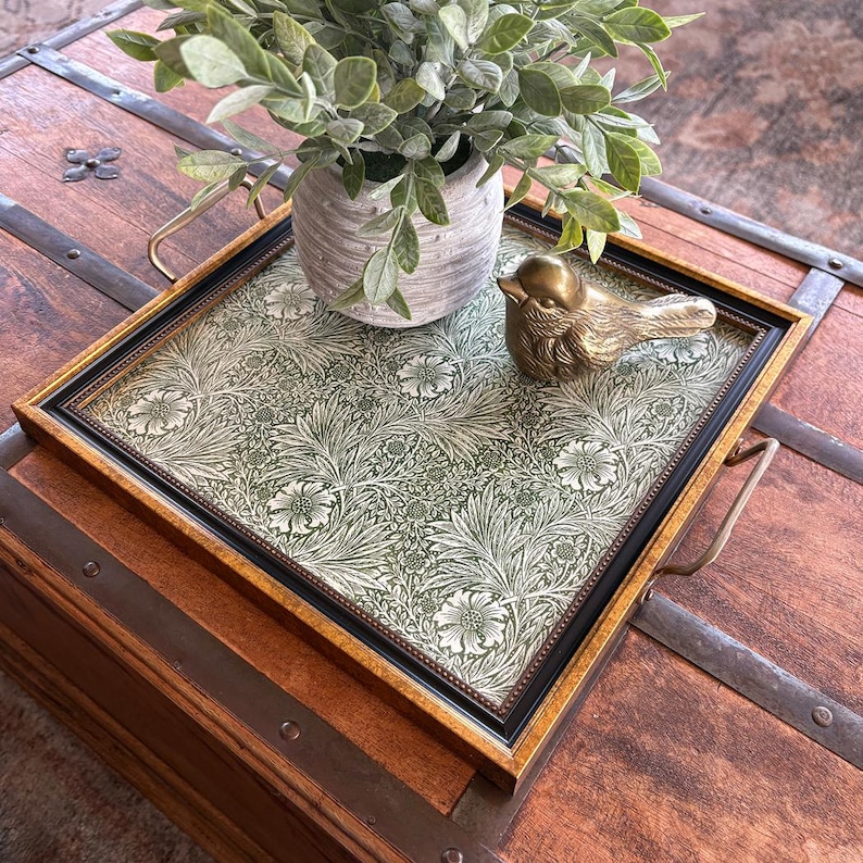 May include: A decorative tray with a floral pattern in shades of green and white, framed in gold and black. A small brass bird figurine and a potted plant with green leaves sit on the tray. The tray has brass handles.