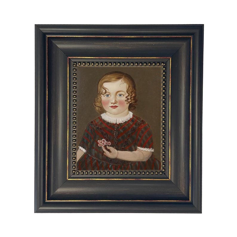 May include: A framed portrait of a young child. The child is wearing a red and black plaid dress and holding a small flower. The portrait is in a black frame with gold accents.