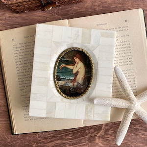May include: A square, white mosaic picture frame with a brass-colored oval frame holding a painting of a mermaid. The painting depicts a red-haired mermaid near the ocean. The frame rests on an open book.