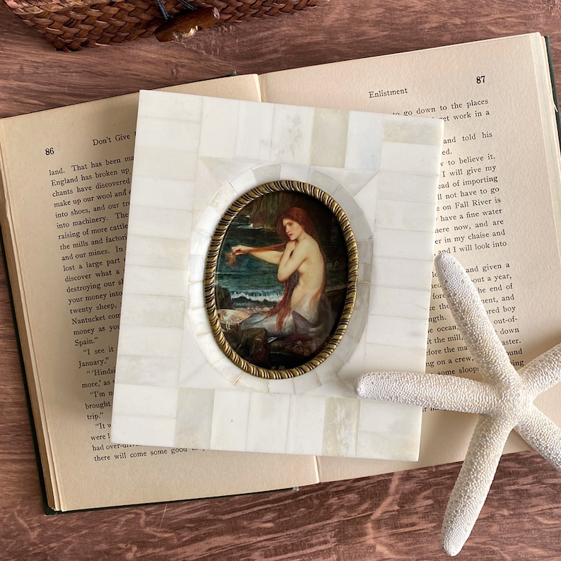 May include: A square, white mosaic picture frame with a brass-colored oval frame holding a painting of a mermaid. The painting depicts a red-haired mermaid near the ocean. The frame rests on an open book.
