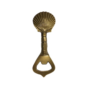 May include: A brass bottle opener with a seashell design. The opener features a detailed, fan-shaped seashell at the top, a cylindrical handle, and a classic bottle-opening mechanism at the bottom. The opener is made of brass and has a shiny finish.