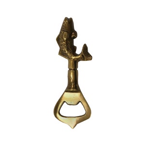 May include: A gold-colored metal bottle opener shaped like a fish. The fish is detailed with scales and has a raised fin. The bottle opener has a classic design with a pointed tip and a circular opening for opening bottles.