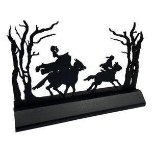 May include: Black silhouette of two figures on horseback riding through a forest. The figures are in the foreground and the trees are in the background. The silhouette is mounted on a black base.