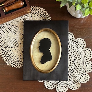 May include: A framed silhouette portrait featuring a black profile against a gold-toned background, encased in an oval brass frame. The frame is set against a black backdrop. The portrait is displayed on a wooden surface with a lace doily.