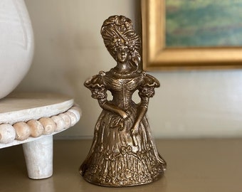 May include: A gold-toned brass bell in the shape of a woman wearing a dress and a hat. The bell has a detailed design and is a vintage style.