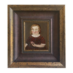 May include: A framed portrait of a child. The child is wearing a red and black plaid dress and holding a small pink flower. The frame is a dark brown with a gold trim.