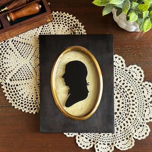 May include: A black and white silhouette of a person's profile in an oval frame with a gold trim. The frame is set in a dark wood frame.