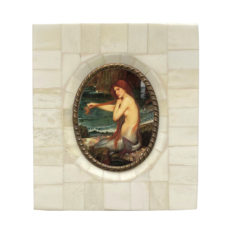 May include: An oval framed artwork featuring a mermaid with long red hair, seated near the ocean. The frame is a gold color, and the background is a square mosaic pattern of white tiles. The mermaid is the focal point of the artwork.