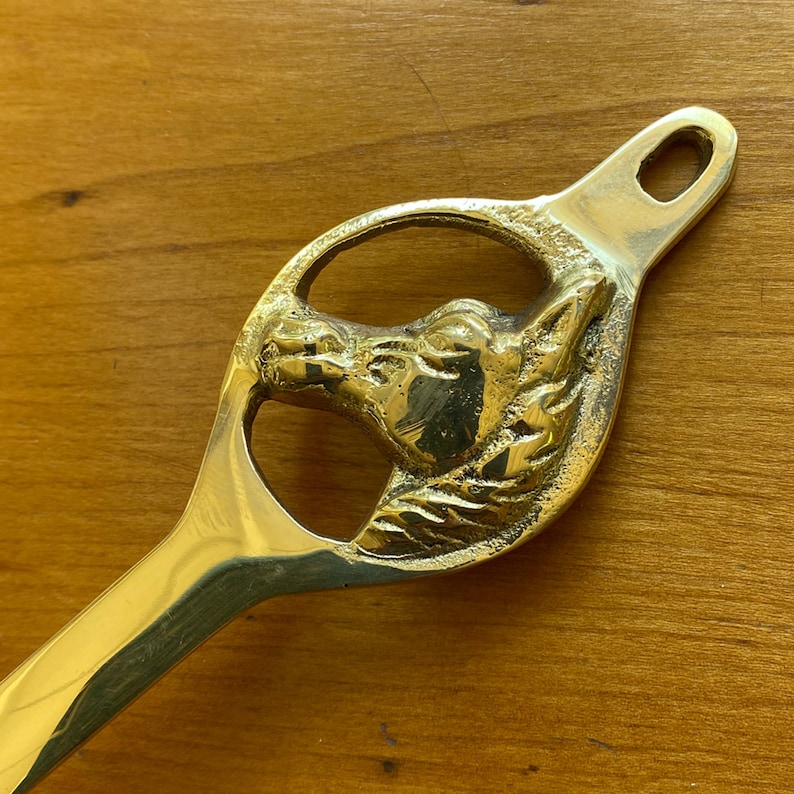 May include: A gold-toned metal horse head doorstop with a detailed design. The horse head is in a circular cutout and the doorstop has a long handle.