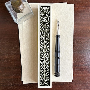 May include: A vintage calligraphy set featuring a black pen with a silver nib, a glass inkwell, and a decorative box with a black and white floral design. The set rests on a stack of parchment paper.