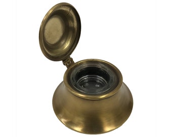 May include: A vintage brass inkwell with a hinged lid. The inkwell has a round, open design with a glass insert. The lid is open, revealing the interior. The inkwell has a classic, antique appearance.