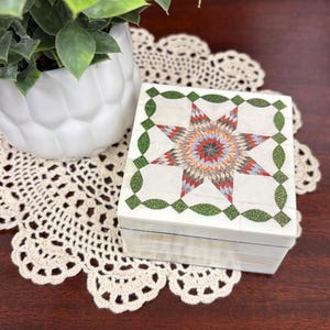 May include: A square, white decorative box with a colorful starburst design on the lid. The starburst features red, orange, blue, and green geometric patterns. The box is trimmed with green leaf-shaped accents.