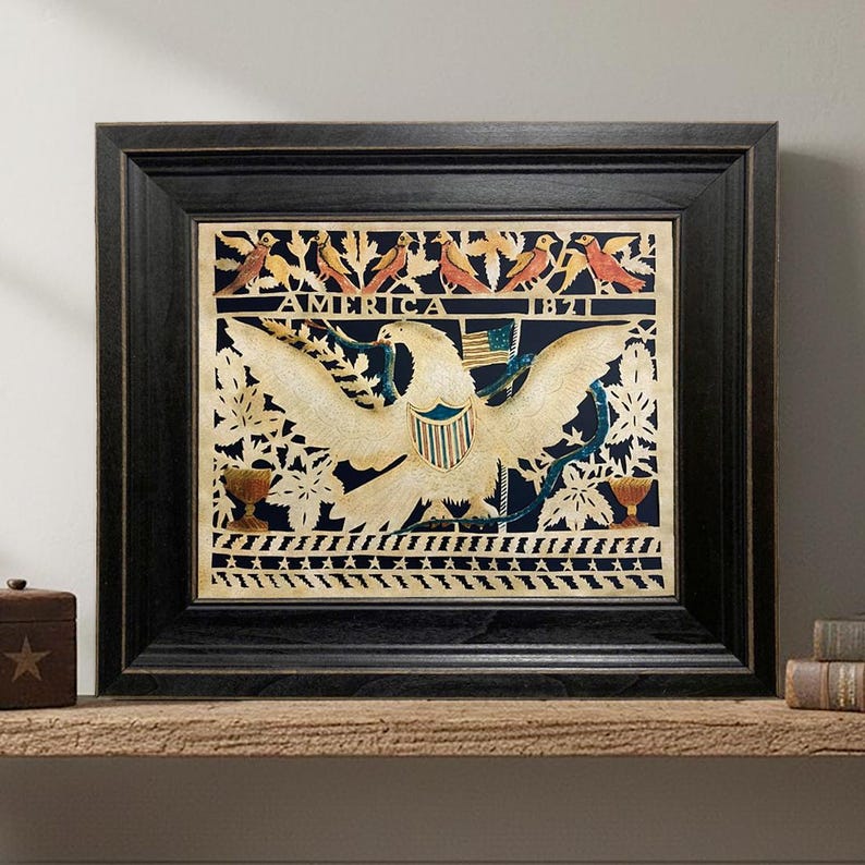 Op de afbeelding: Een ingelijst papierknipkunstwerk met een centrale witte adelaar met gespreide vleugels, een schild en een kleine vlag. De tekst AMERICA 1821 staat gecentreerd boven de adelaar. Het ontwerp bevat vogels, bladeren en twee urnen op een donkerblauwe achtergrond.