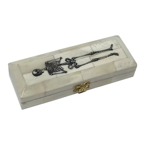 May include: A white bone inlay box with a skeleton design on the lid. The box has a gold metal clasp.