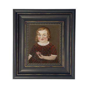 May include: A framed portrait of a young child. The child is wearing a red and black plaid dress and holding a small flower. The portrait is in a black frame with gold accents.