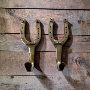 May include: Two brass horseshoe-shaped wall hooks. Each hook has a curved top with drilled holes and a downward-facing hook. The hooks are mounted on a rustic wooden background, creating a vintage aesthetic.