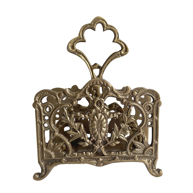 May include: Ornate brass letter holder with a decorative floral and insect design. The vintage desk organizer features a filigree pattern and a curved handle at the top. The brass has a warm, aged patina.