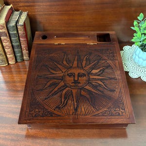 Könnte beinhalten: Eine dunkelbraune Holzschreibkiste mit einem eingravierten Sonnendesign auf dem Deckel. Die Kiste hat einen Klappdeckel und ein kleines Fach. Das Sonnendesign befindet sich zentral, umgeben von einem dekorativen Rand.