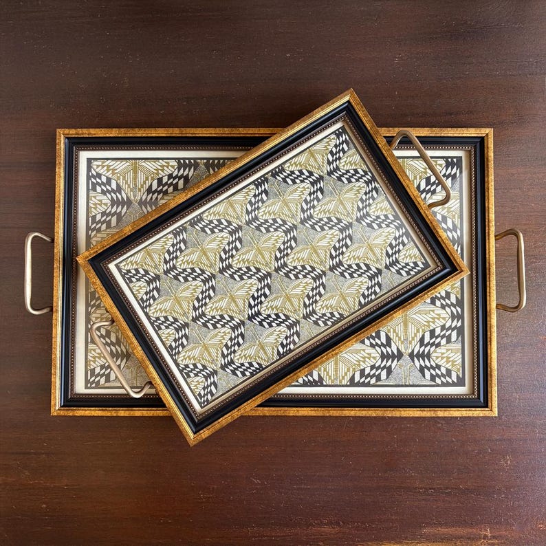 May include: Two decorative serving trays with gold frames and handles. The trays feature a black, white, and gold geometric pattern. One tray is placed on top of the other, creating a layered effect. The trays are on a dark brown wooden surface.