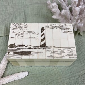 May include: A rectangular off-white box constructed from small, joined panels. The lid features a dark brown etched illustration of a striped lighthouse, a small boat on the water, and coastal buildings under a sky with clouds.