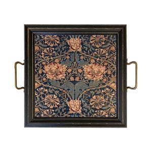 May include: A square serving tray with a black frame and two brass-colored metal handles. The center features a dark blue fabric inlay with a repeating pattern of light pink flowers, tan petals, and blue-green foliage arranged in a symmetrical design.