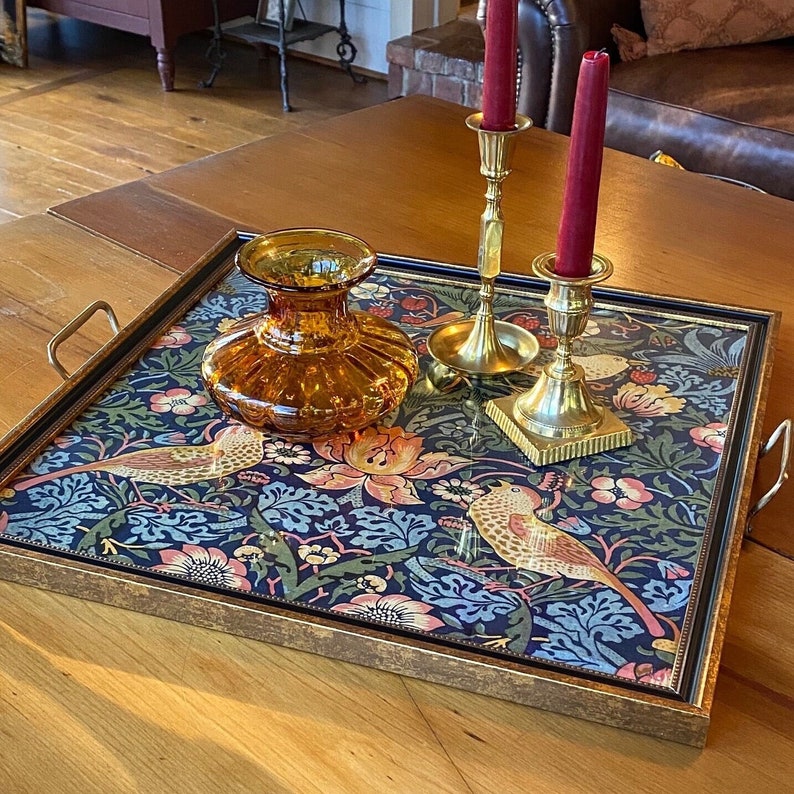 K&ouml;nnte beinhalten: Ein dekoratives Tablett mit Blumen- und Vogelmuster, mit einem goldfarbenen Rahmen und Griffen. Eine bernsteinfarbene Vase und zwei goldene Kerzenst&auml;nder mit roten Kerzen stehen auf dem Tablett und schaffen eine Vintage-&Auml;sthetik.