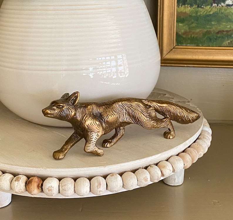May include: A gold-toned metal fox figurine on a white wooden pedestal with a beaded edge. The fox is standing on all four paws with its tail curled behind it.