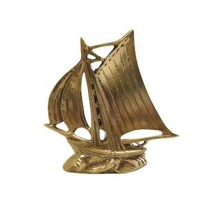 May include: A small, gold-colored metal sailboat figurine. The sailboat has a single mast and a sail with detailed lines. The figurine is sitting on a white background.