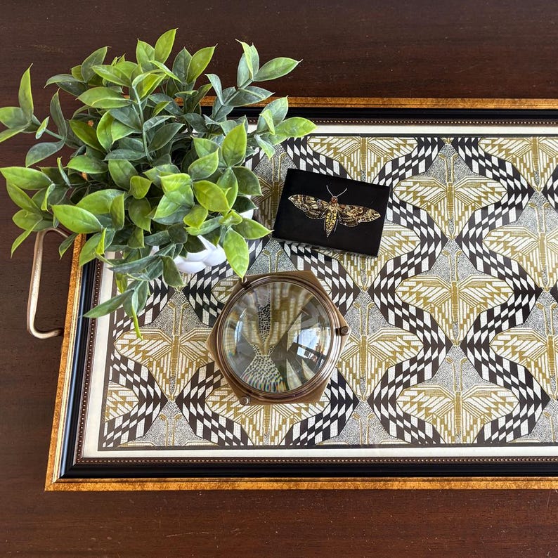 May include: A decorative tray with a geometric pattern in black, white, and gold. A small black box with a gold moth design, a magnifying glass, and a potted plant are on the tray. The tray has a black frame with gold trim.