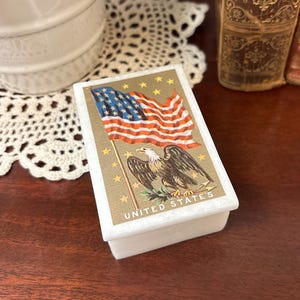 May include: A small, rectangular white box with a decorative top featuring an American flag and an eagle illustration. The flag is red, white, and blue, and the eagle is brown and white. The words "UNITED STATES" are printed below the eagle.