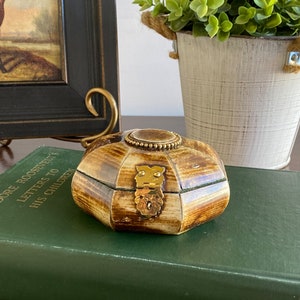 Op de afbeelding: Een kleine, achthoekige houten doos met een messing sluiting en decoratieve versieringen. De doos is gesloten en heeft een bruin en beige houtnerfpatroon.