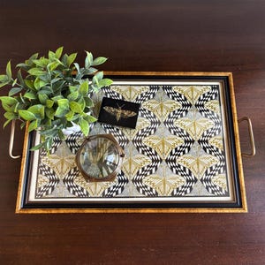 May include: Decorative serving tray with a black frame and gold handles. The tray features a patterned design in black, white, and gold. A small black card with a gold moth design, a magnifying glass, and a small plant are on the tray.