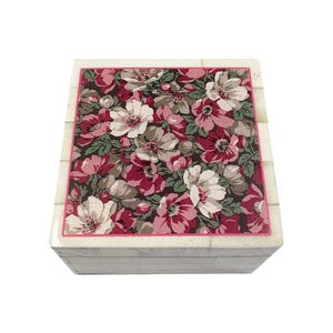 May include: A square decorative box with a floral pattern on the lid. The lid features a pink border and a design of white, pink, and burgundy flowers on a dark background. The box has a light-colored, textured exterior.