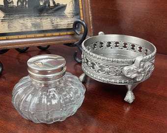May include: A clear glass inkwell with a ribbed body and a silver-toned metal cap, positioned next to a circular silver-toned metal stand featuring an intricate openwork lattice pattern, beaded edges, and three decorative claw-style feet.