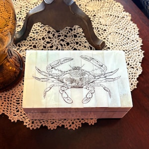 May include: A rectangular, off-white box with a detailed crab illustration on the lid. The crab is depicted in black ink, and the box has a light, pearlescent finish. The box is resting on a cream-colored, crocheted doily.