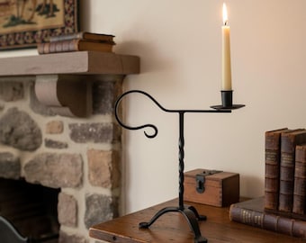 May include: Black wrought iron candle holder with a lit cream-coloured candle. The candle holder has a twisted stem and a curved handle. The candle holder is on a wooden table with books and a small wooden box.