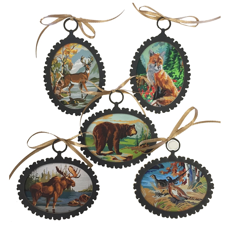 May include: Five oval-shaped ornaments with black metal frames and gold ribbon hangers. Each ornament features a different woodland animal illustration: a deer, a fox, a bear, a moose, and a pair of birds.