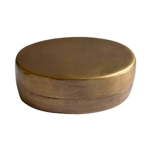 May include: A small, oval-shaped container with a lid, crafted from a solid, brushed brass-toned metal. The object features a smooth, matte finish with subtle horizontal graining across the surface, presenting a simple, rounded, and compact form.