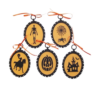 May include: Five black and orange Halloween ornaments with different spooky designs. The ornaments are shaped like ovals with a black scalloped border and a gold background. The designs include a skeleton, a spider web, a pumpkin, a haunted house, and a rider on a horse.
