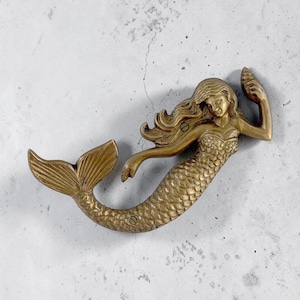 May include: A brass-colored metal mermaid wall hook or door handle featuring a detailed scale pattern on the tail, flowing hair, and one arm raised holding a seashell. The object includes two visible screw holes for mounting purposes.