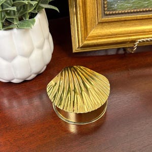 May include: A gold-coloured seashell-shaped trinket box. The lid is textured with radiating lines, and the base is a simple cylinder. The box sits on a dark wood surface, with a white decorative pot and a framed picture in the background.